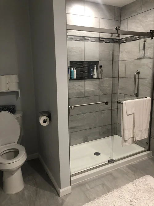 Bathroom with a glass-enclosed shower featuring gray tile walls and a built-in shelf with toiletries