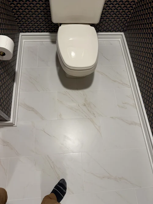 White ceramic toilet with a closed lid in a bathroom with marble-patterned floor tiles and dark patterned wallpaper
